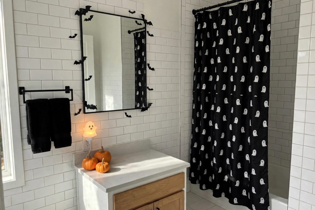 Halloween bathroom decor in a guest bathroom with a black ghost shower curtain, black towels, mirror bat decals and a ghost night light