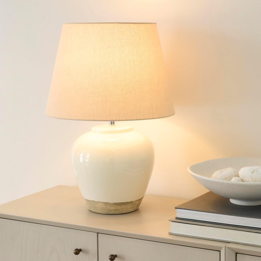Nourison ceramic table lamp lit on a light wood console, warm light through the beige shade